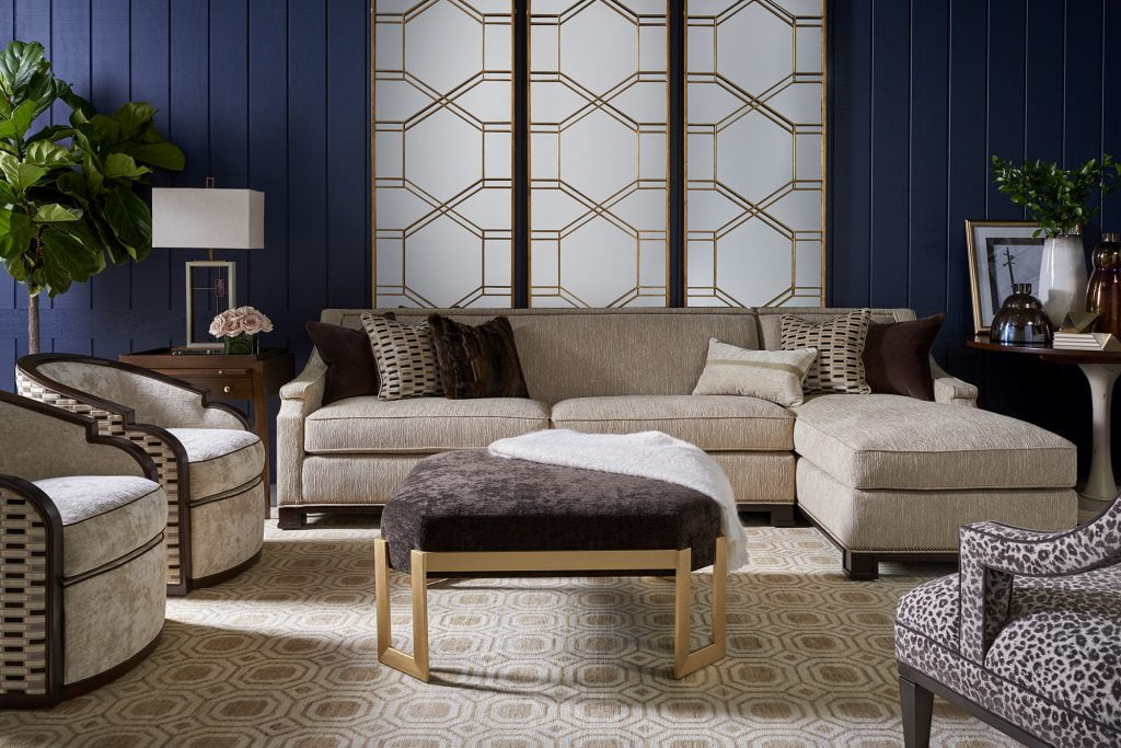 studio shot of living room set from Wesley Hall. Navy blue walls with beige sectional and brown and gold ottoman