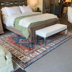 wooden bed frame with a green comforter and multicolored rug and white cushioned bench