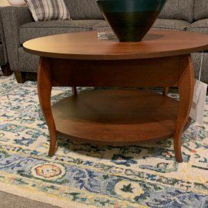 Round wooden coffee table in chestnut on top of a blue, yellow, cream, and green patterned rug.