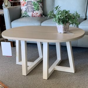 Wooden beige table with white geometric legs. Flower in vase on table top with blue couch in background.